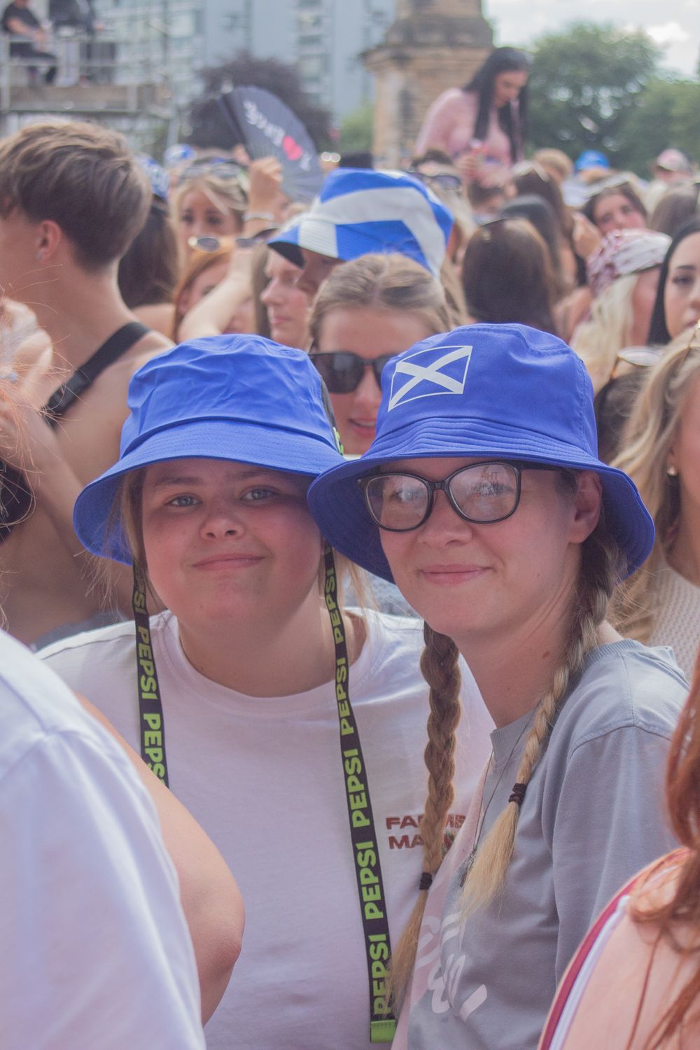 Two smiling women wearing blue Scottish flag bucket hats at a crowded outdoor music festival.