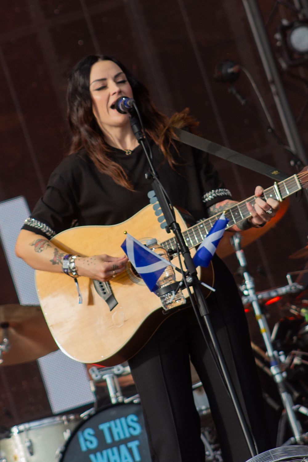 Woman in black outfit plays acoustic guitar with Scottish flag on stage at live concert.