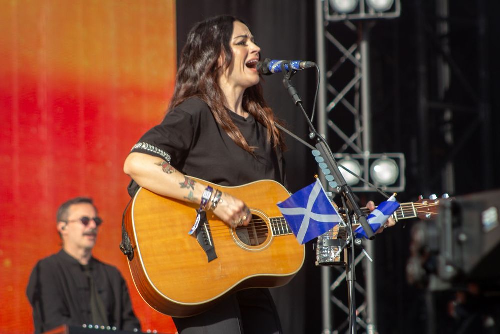 Female guitarist with tattoos performs on outdoor stage with acoustic guitar and Scottish flag.