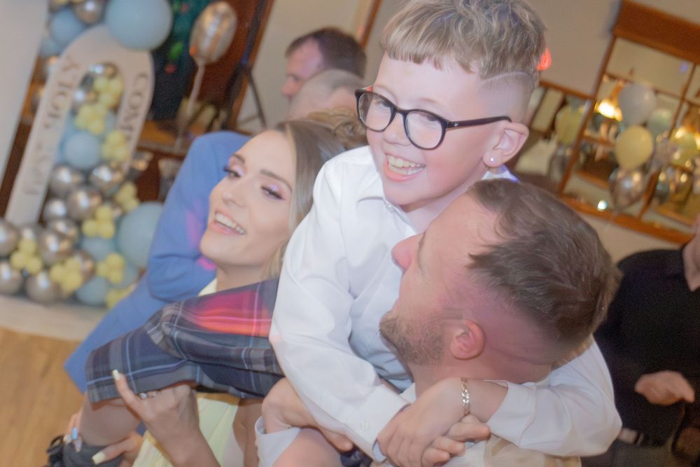 Boy in glasses and kilt laughs while being embraced by family at First Communion celebration.