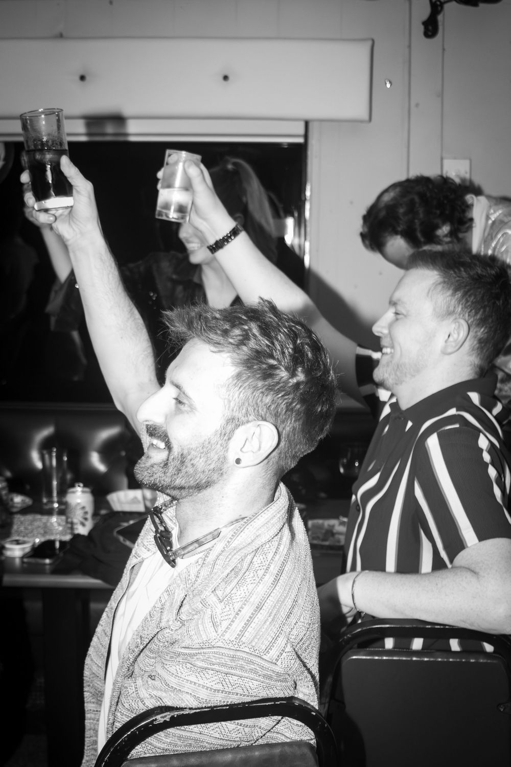 Black and white photo of bearded man raising a drink at a lively party while friend smiles beside him.