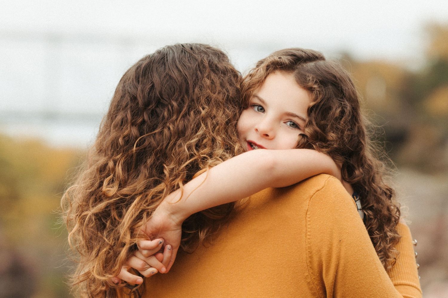 Small girl with curly hair smiling while hugging her mom and looking over her shoulder.