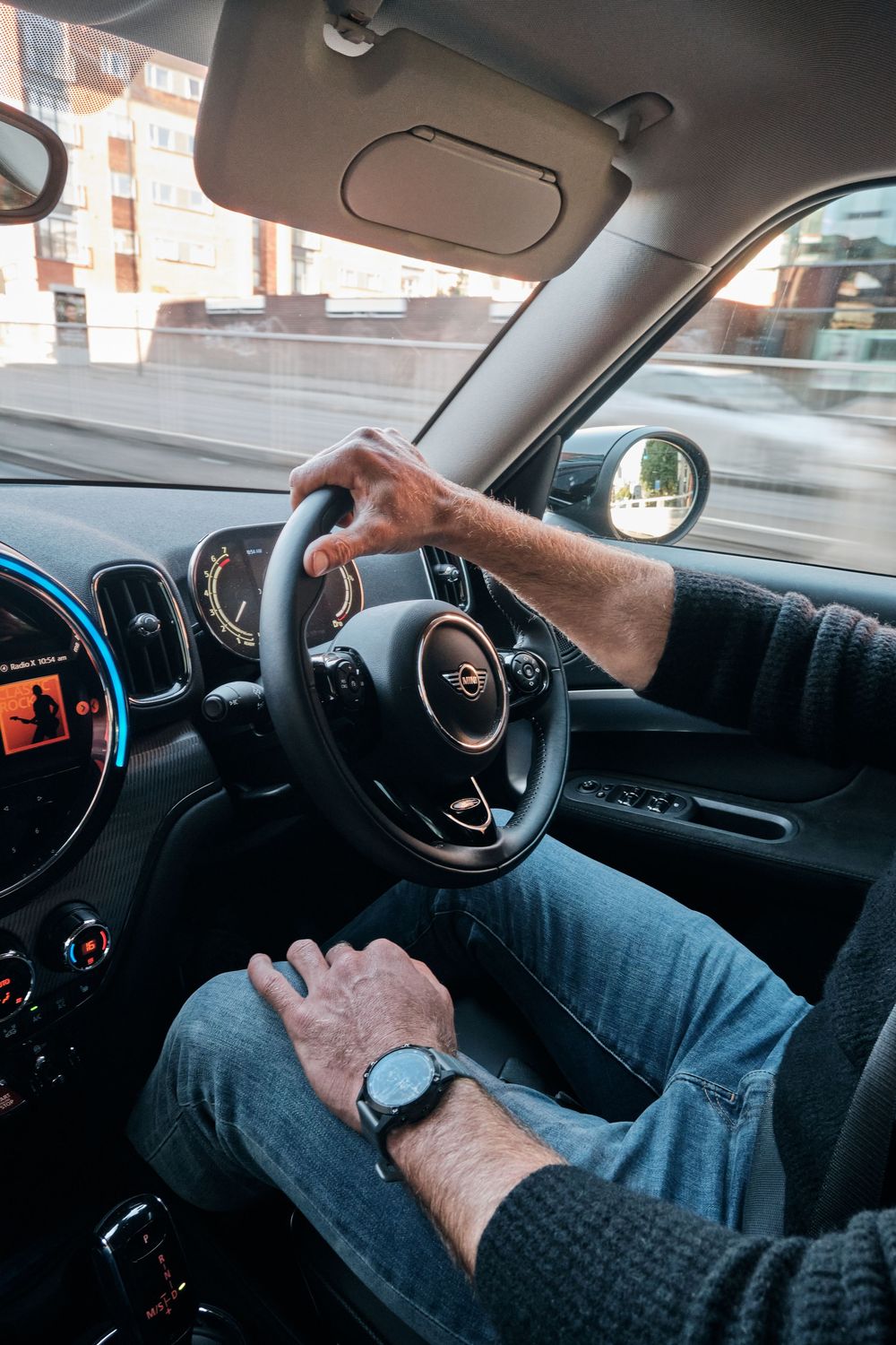 Man driving a MINI Cooper car, hands on steering wheel, wearing a watch, blue jeans, modern dashboard visible.