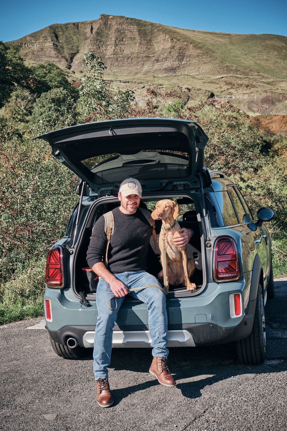 Man and brown dog sitting in open car trunk with scenic green hills in background on sunny day.
