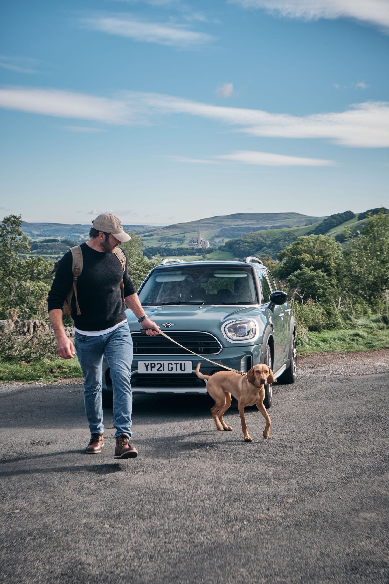 Man walking a golden dog on a leash beside a MINI Countryman car on a rural road with scenic hills.