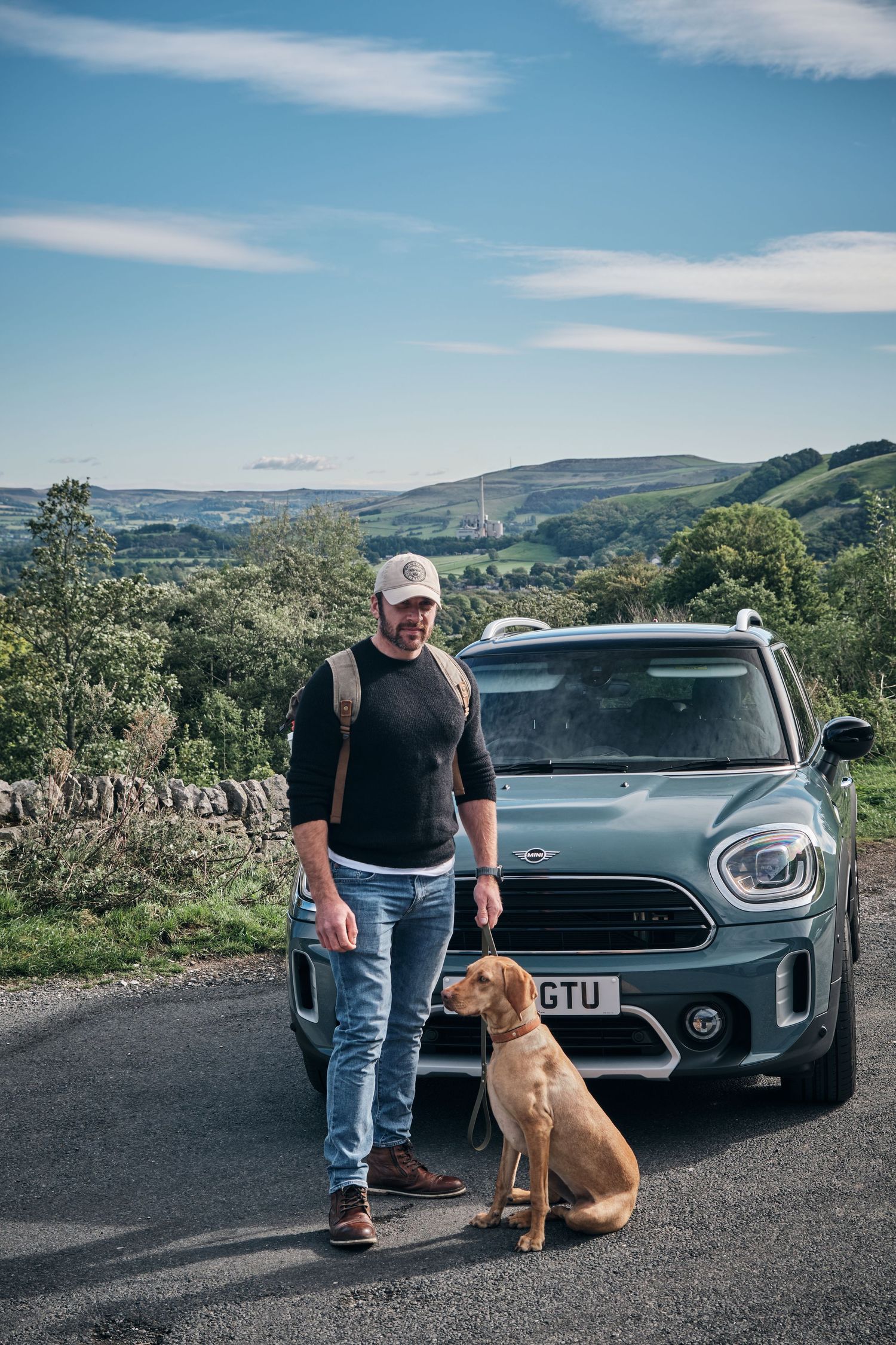 Man in cap and backpack stands with brown dog beside a green MINI Countryman on a scenic rural road.