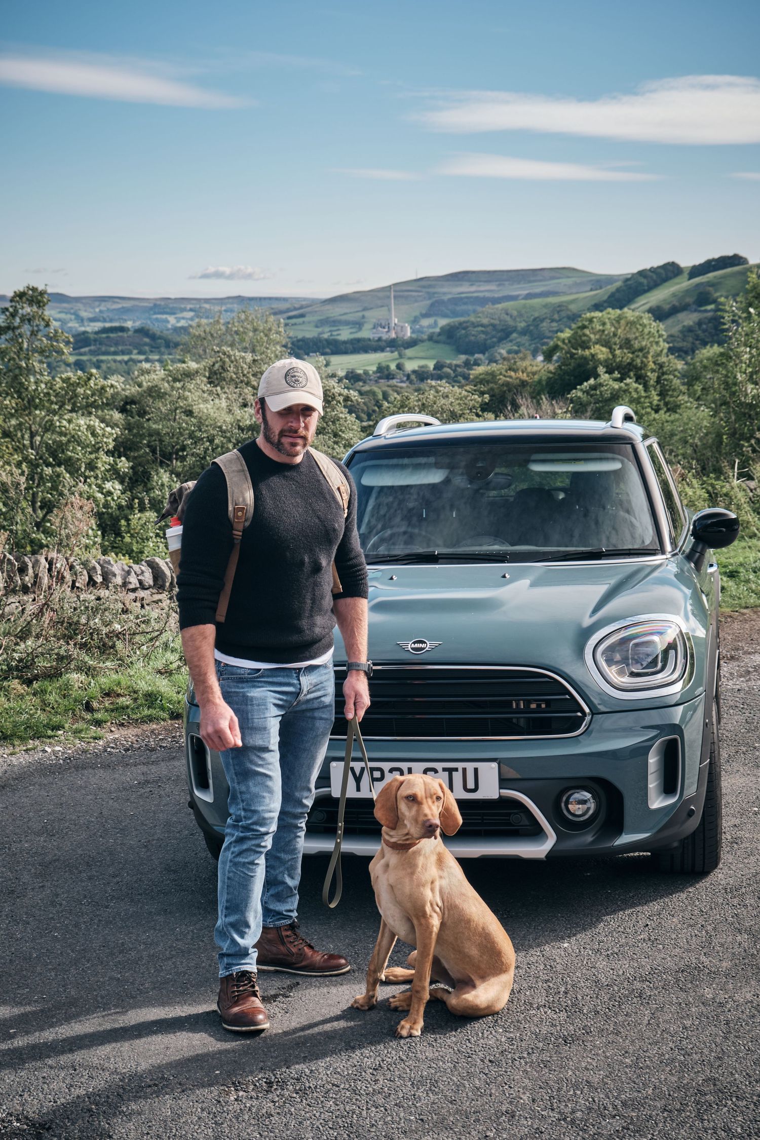 Man in cap and backpack stands with a brown dog beside a MINI Countryman on a scenic rural road with rolling hills.