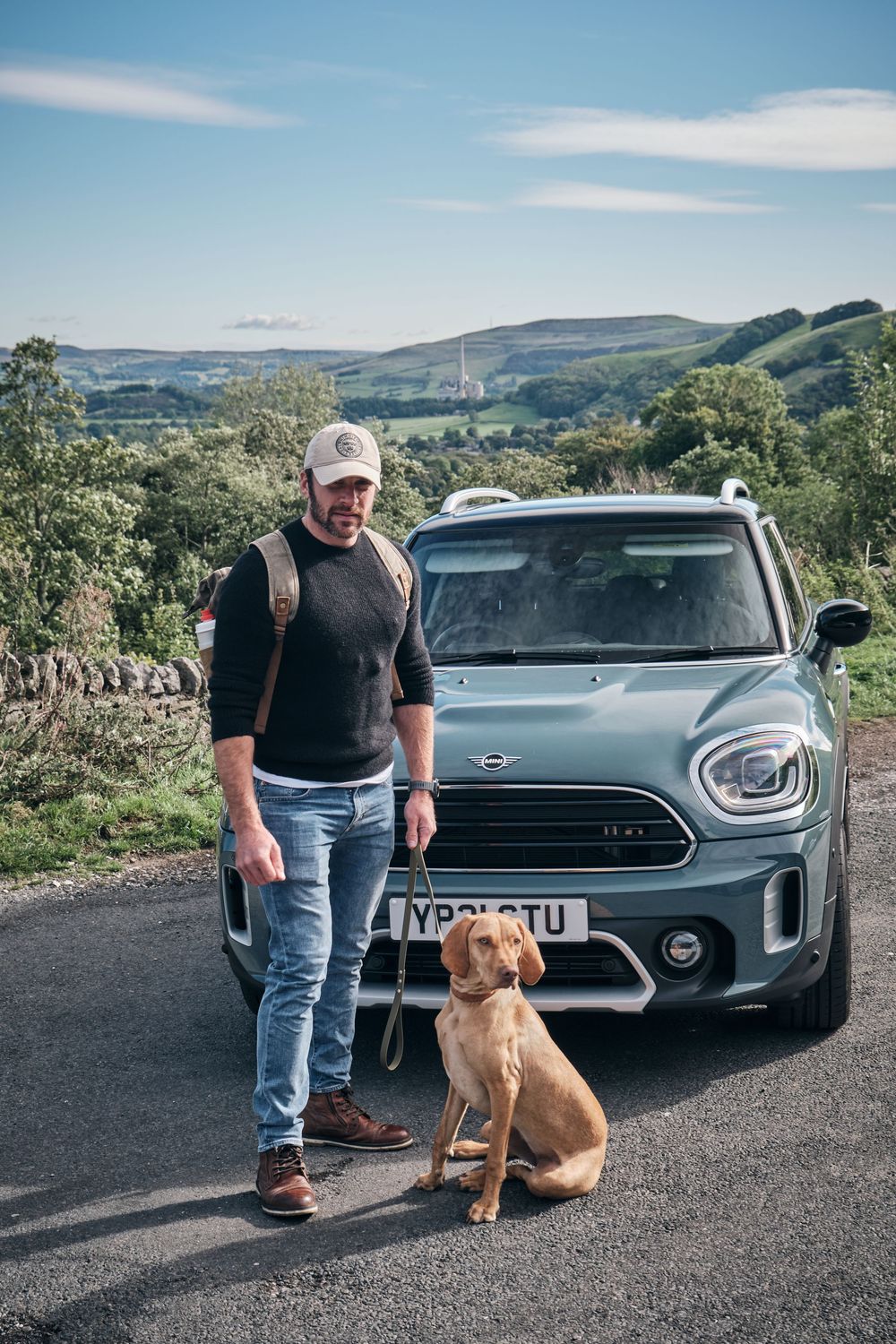 Man in cap and backpack stands with a brown dog beside a MINI Countryman on a scenic rural road with rolling hills.