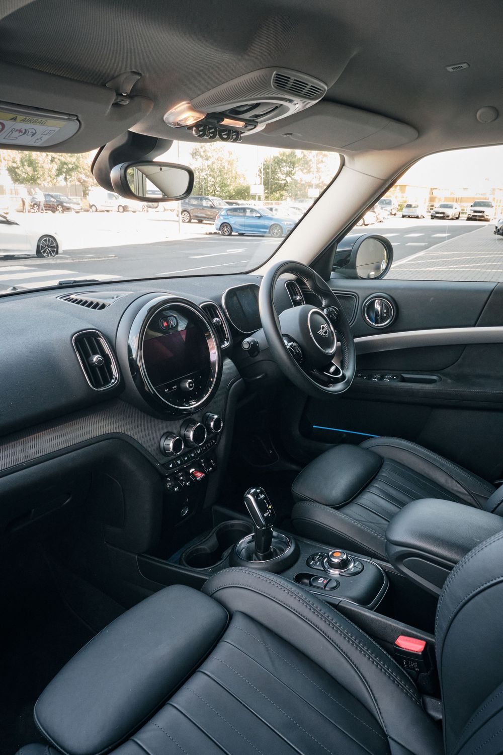 Interior view of a MINI Countryman with black leather seats, circular infotainment screen, and manual gearshift in a parking lot.