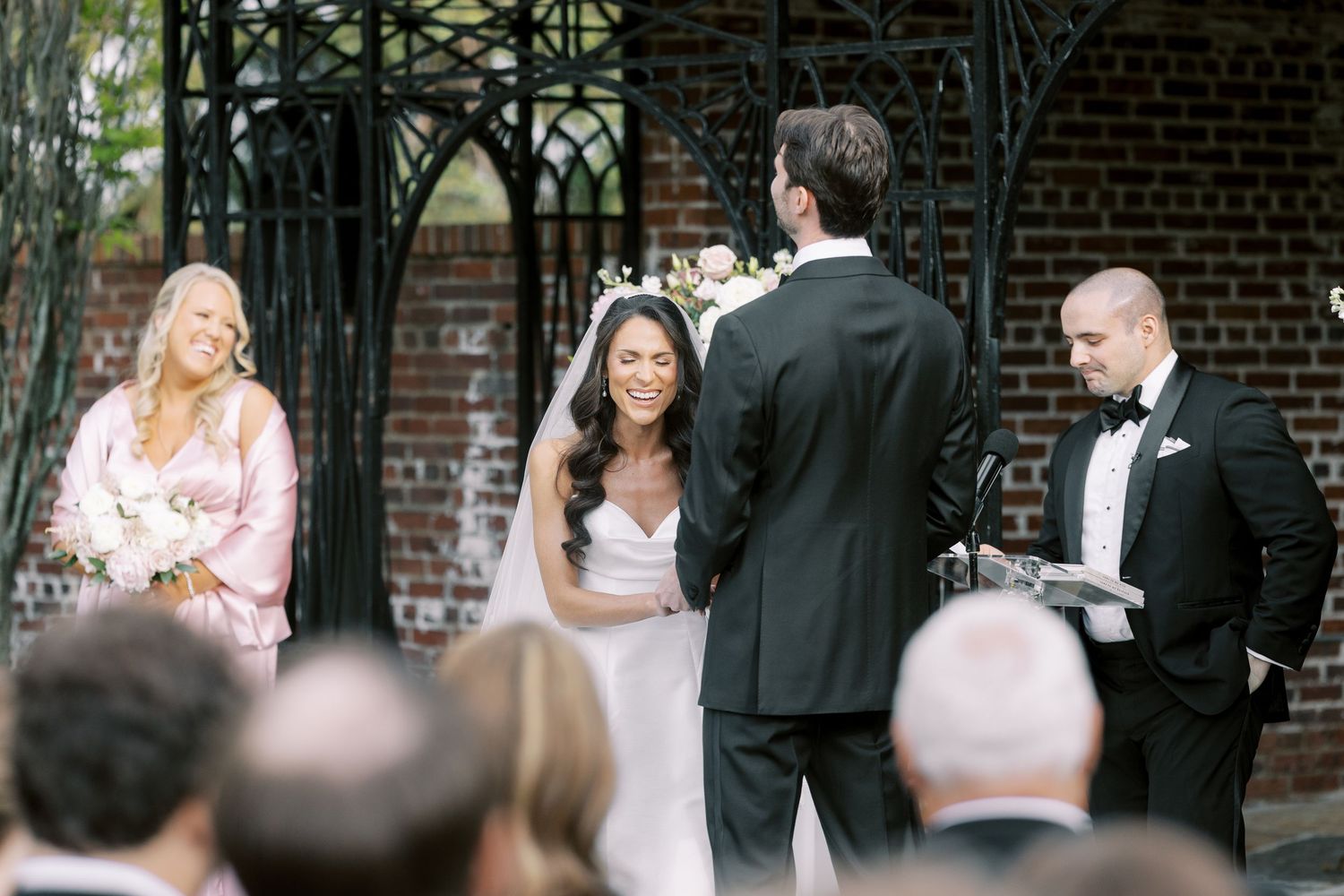 Outdoor wedding ceremony with bride in white gown, groom in tuxedo, officiant, and bridesmaid in pink dress.