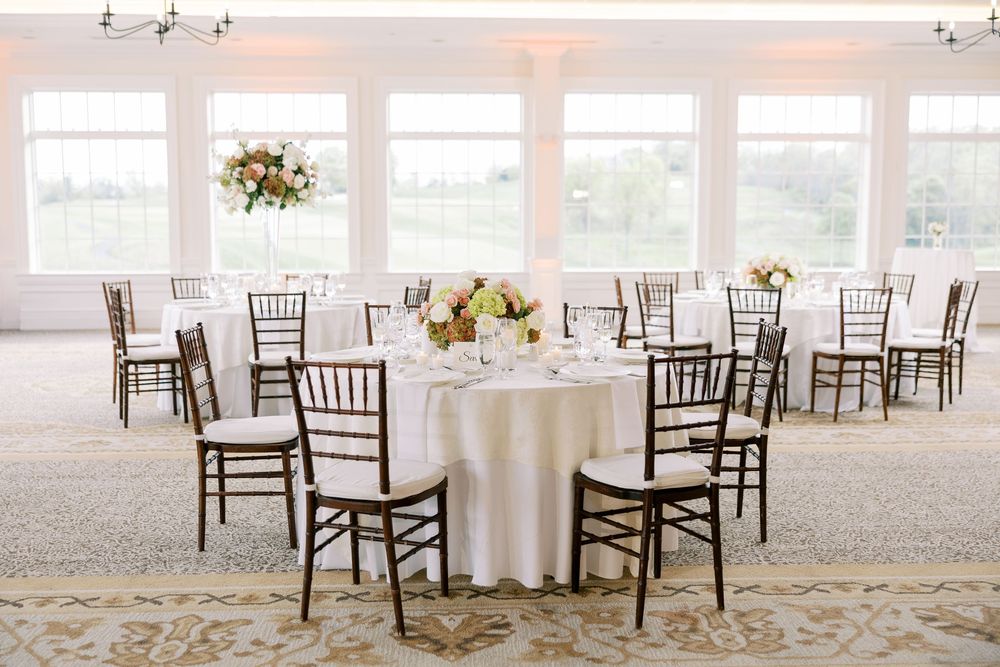 Elegant wedding reception hall with round tables, white linens, floral centerpieces, and dark Chiavari chairs by large windows.