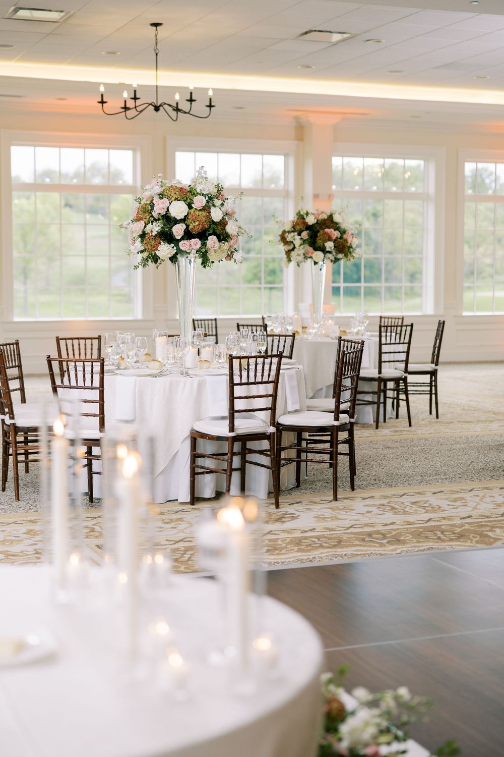 Bright wedding reception hall with tall floral centerpieces, chiavari chairs, and glowing candles.