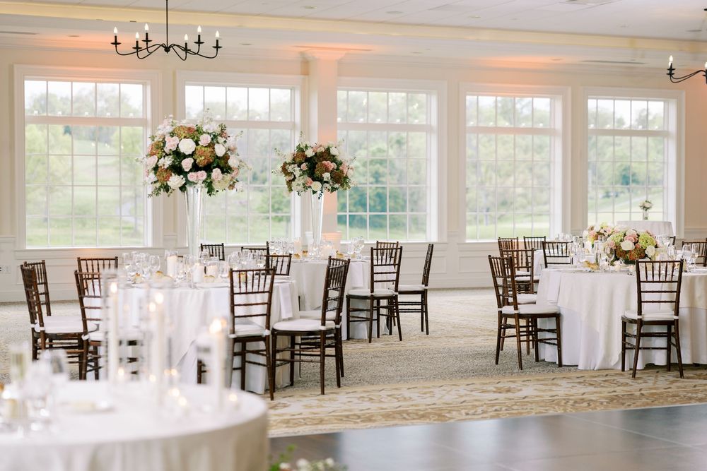 Elegant wedding reception hall with white tablecloths, dark wood chairs, tall floral centerpieces, and large windows with natural light.