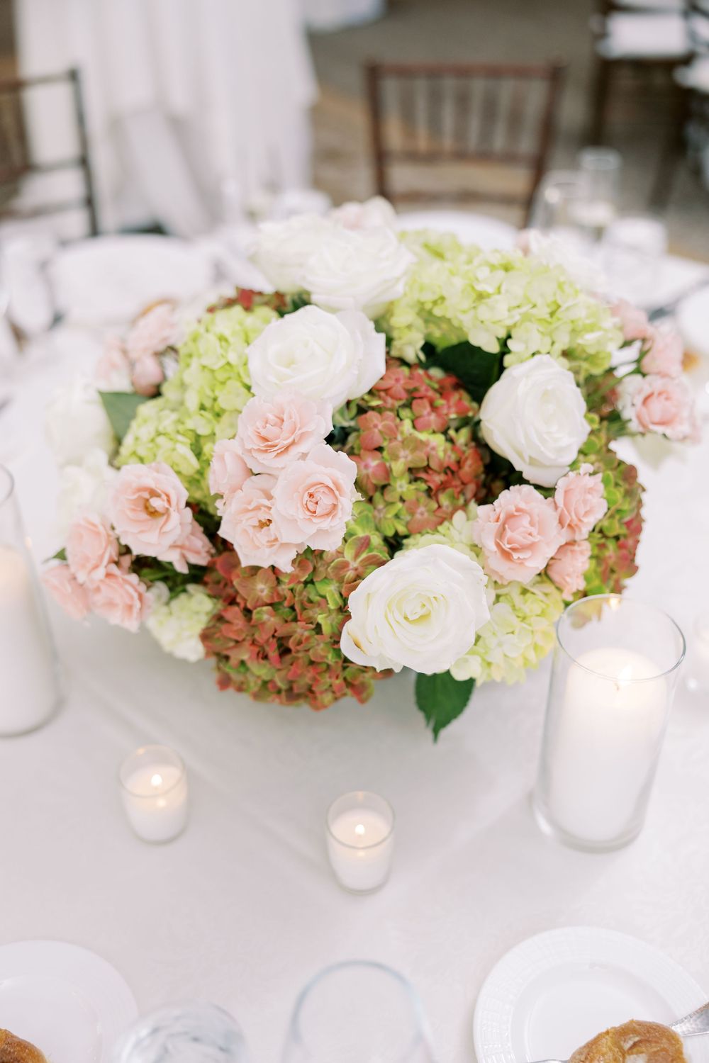 Elegant wedding centerpiece with pink roses, white lisianthus, and green hydrangeas in a bowl vase, surrounded by candles.