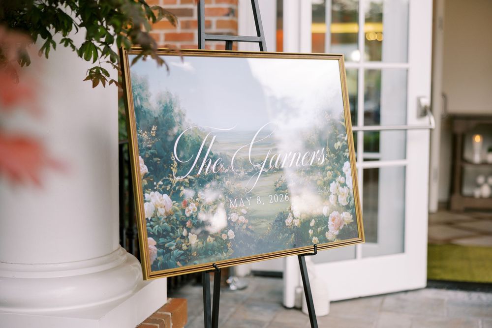 Elegant gold-framed wedding welcome sign on easel with floral background displaying The Garners in white script calligraphy.