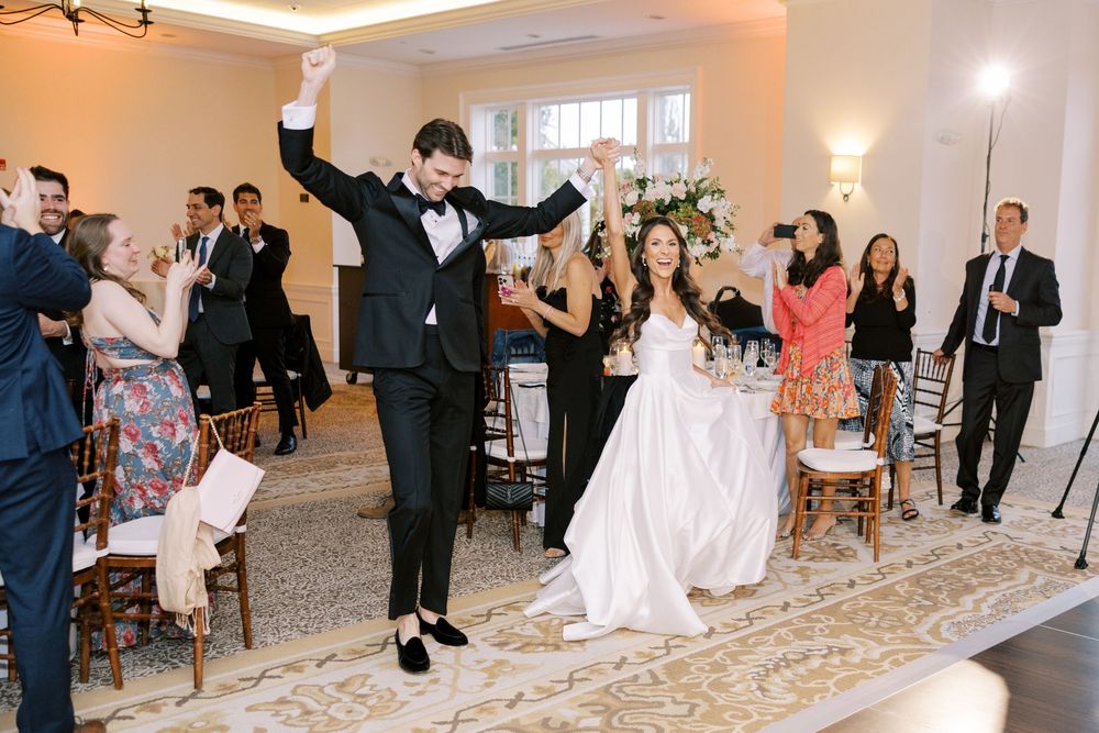 Groom raises arm in celebration as bride sits smiling during wedding reception entrance with guests applauding around them.