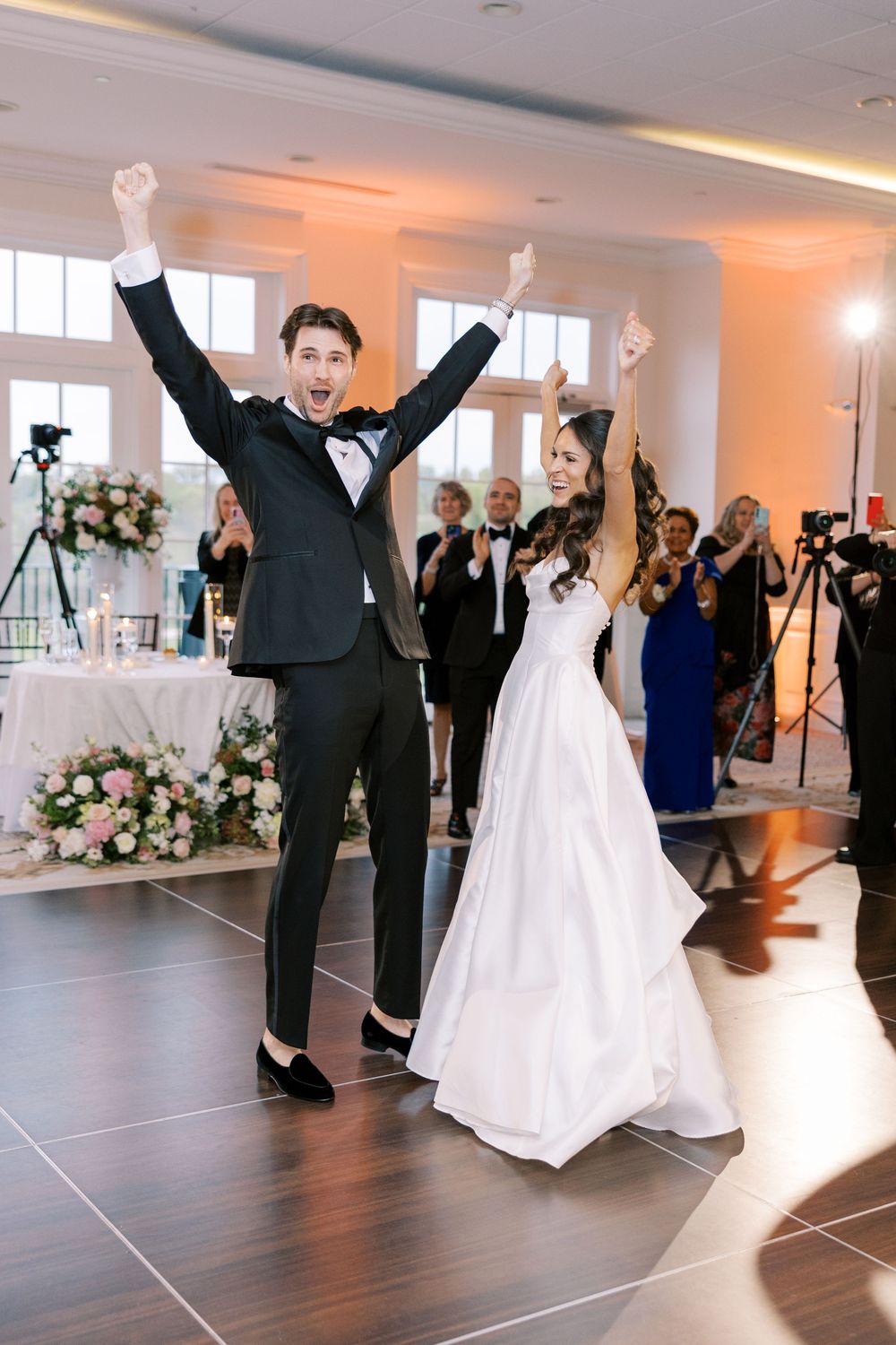 Joyful bride and groom celebrate their first dance, arms raised in excitement, at an elegant wedding reception.