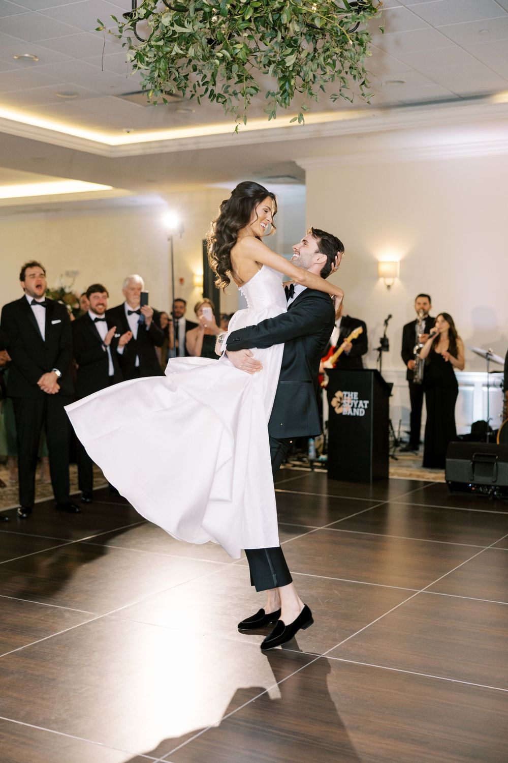 Groom lifts bride in white gown during first dance at elegant wedding reception with live band performing.