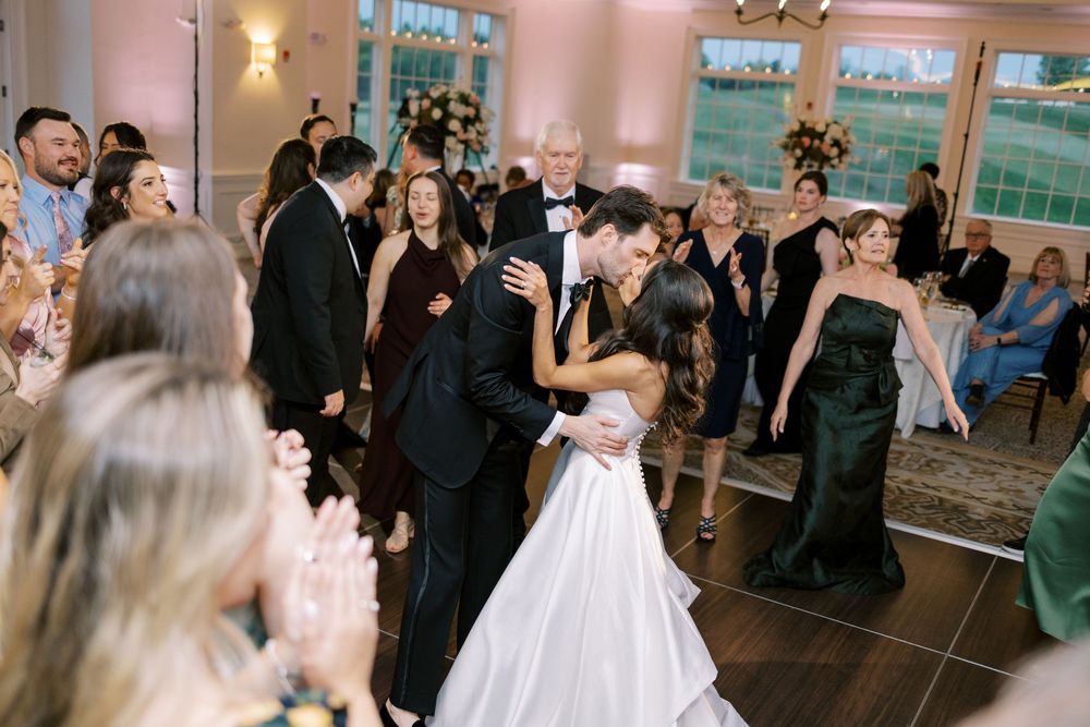 Bride and groom share a romantic first dance kiss surrounded by cheering wedding guests in an elegant reception venue.