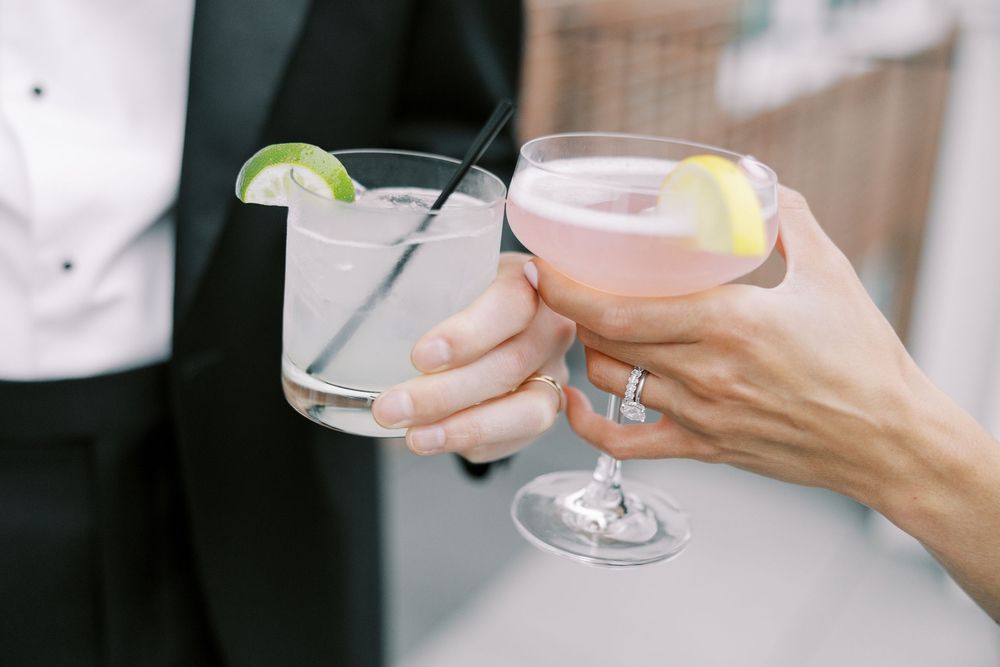 Two hands clinking cocktail glasses with citrus garnishes at a wedding celebration, bride wearing diamond ring.