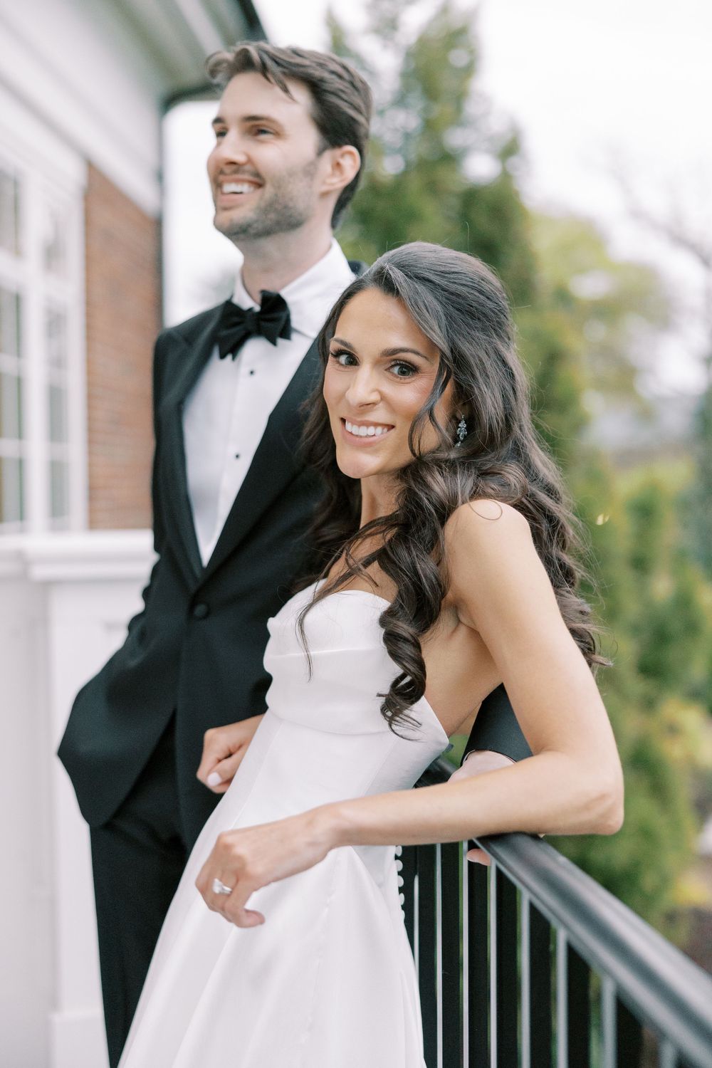 Smiling bride in strapless white gown stands with groom on balcony at elegant wedding venue.