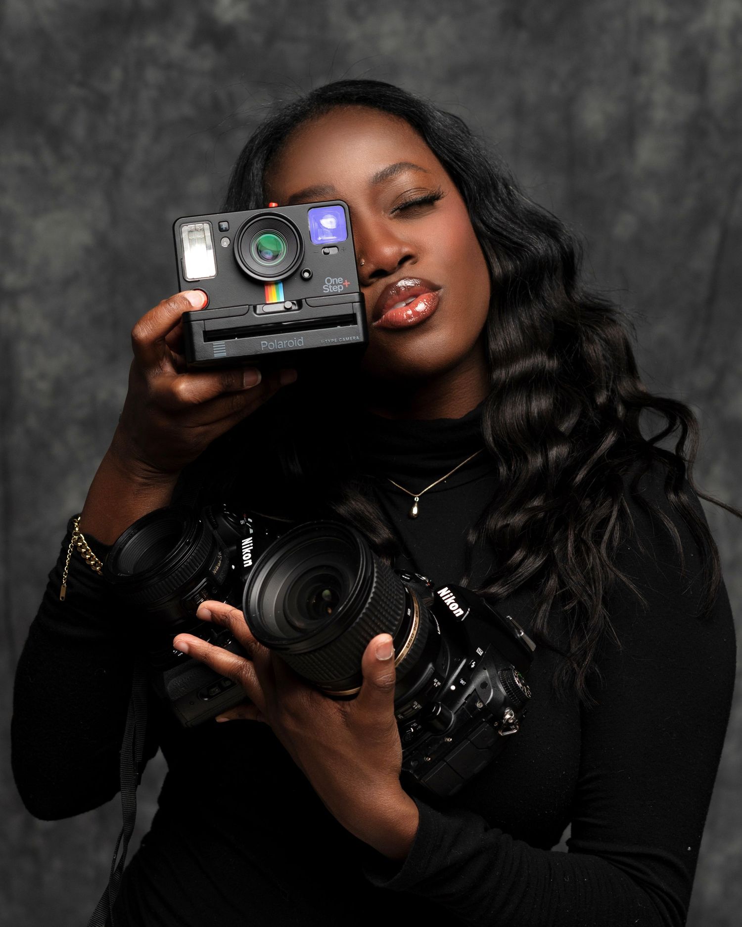 A young woman holds a Polaroid camera up to her face and a Nikon DSLR in her other hand against a gray backdrop.