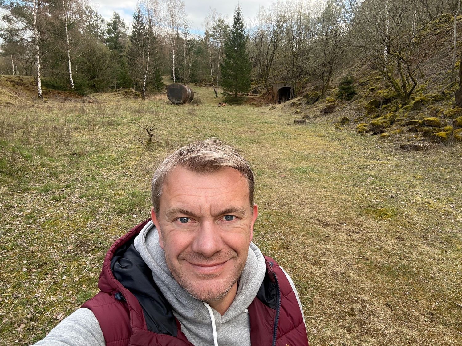 Man taking a selfie in a scenic forest clearing with trees and a stone tunnel in background.