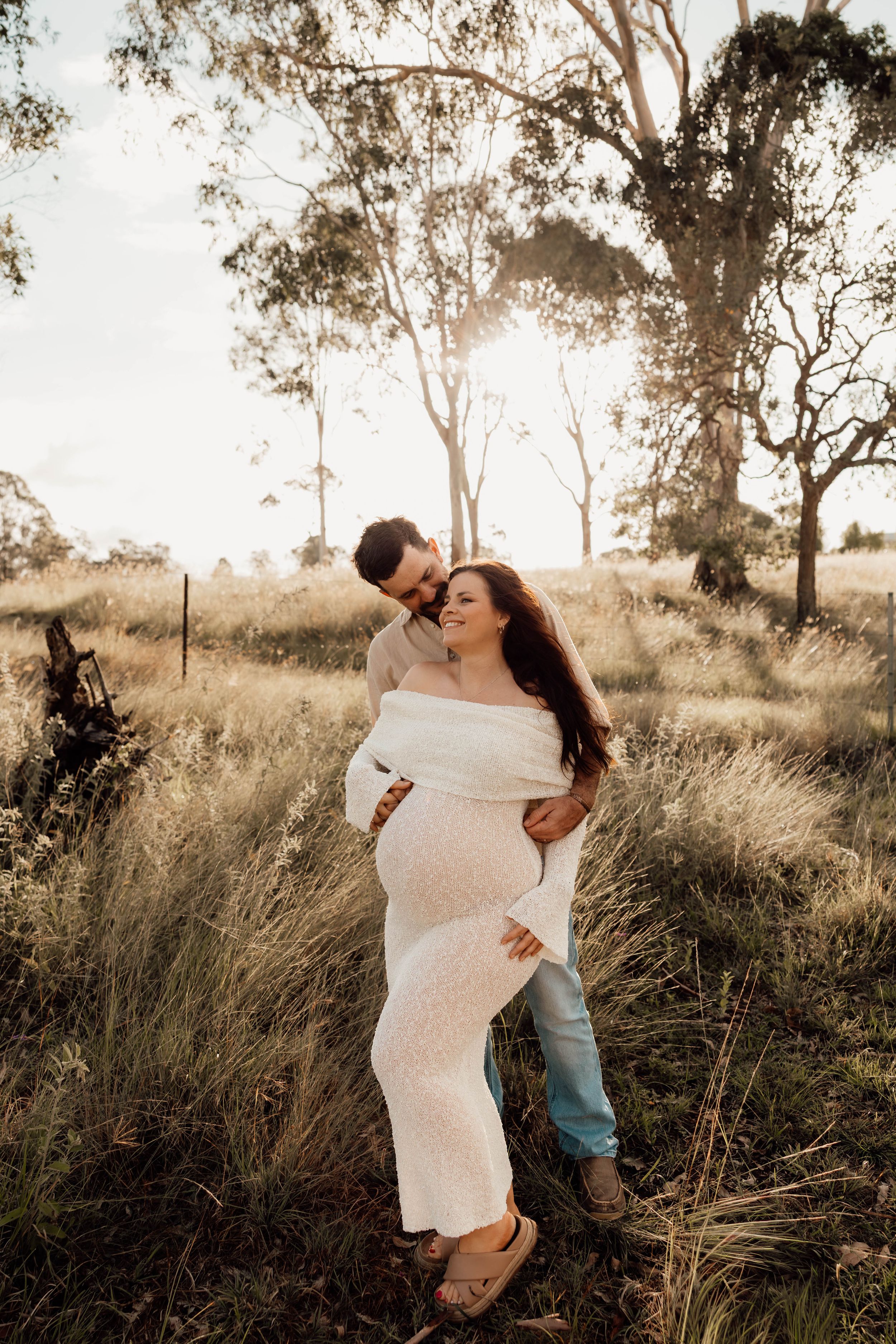 Couple share a tender moment in golden hour light, man embracing pregnant woman in white dress in rural Australian field.
