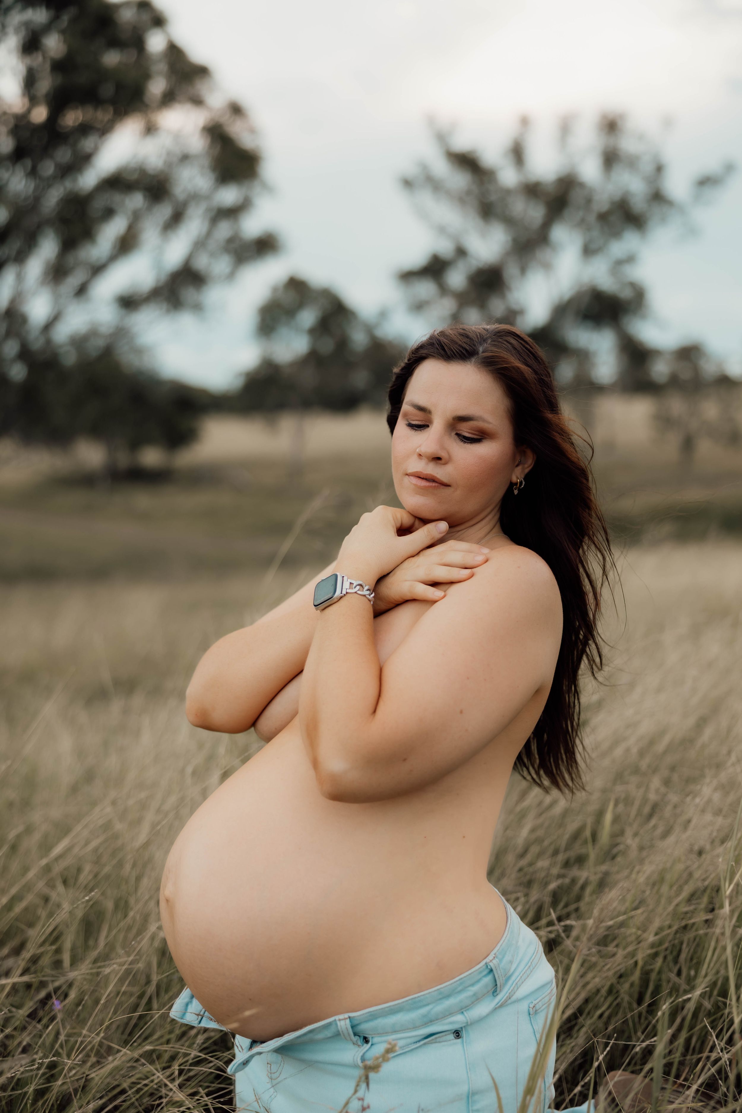 Topless pregnant woman with dark hair poses in a field, eyes closed, hands crossed over chest.