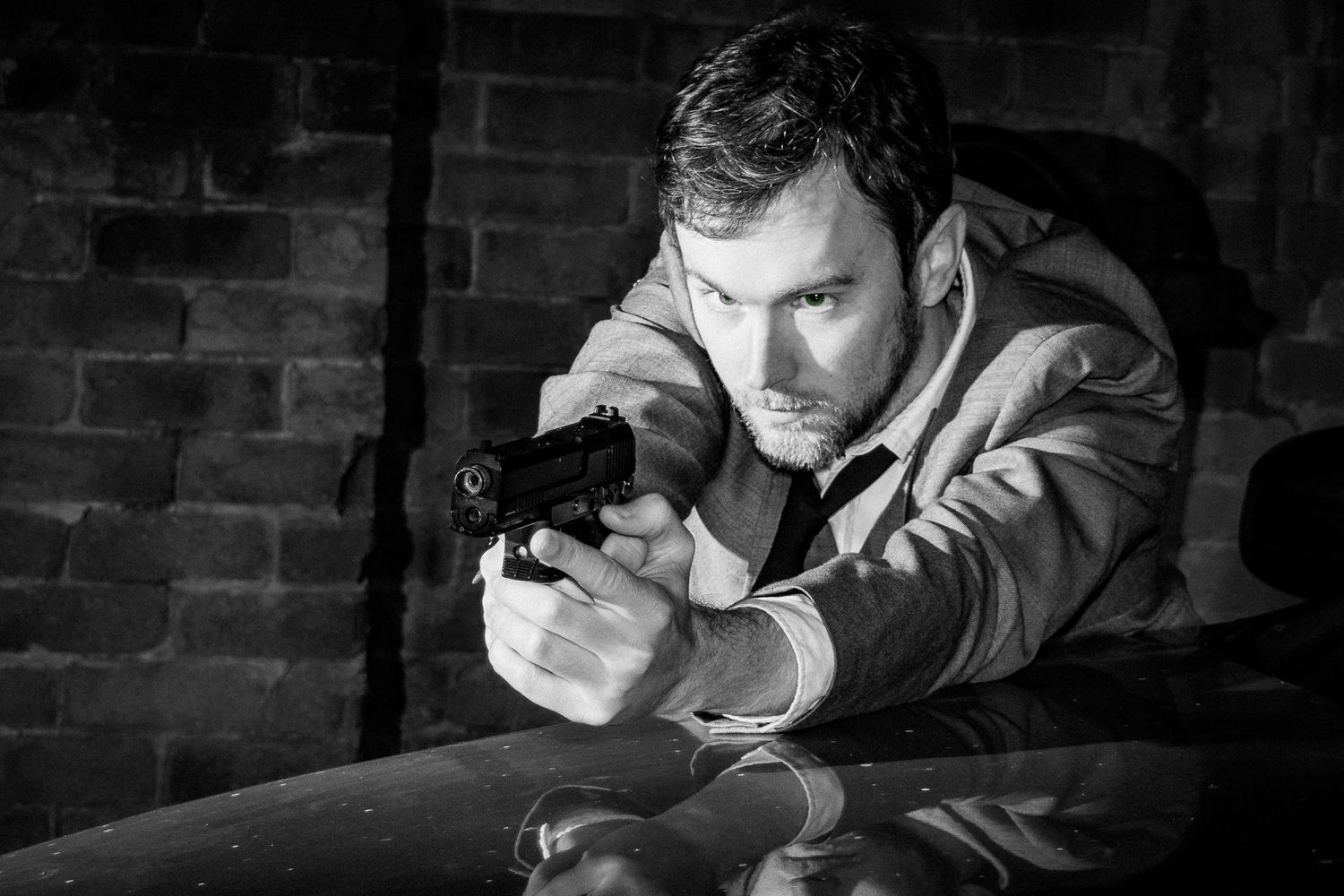 Black and white photo of a man in a suit aiming a handgun, dramatic noir style against a brick wall.