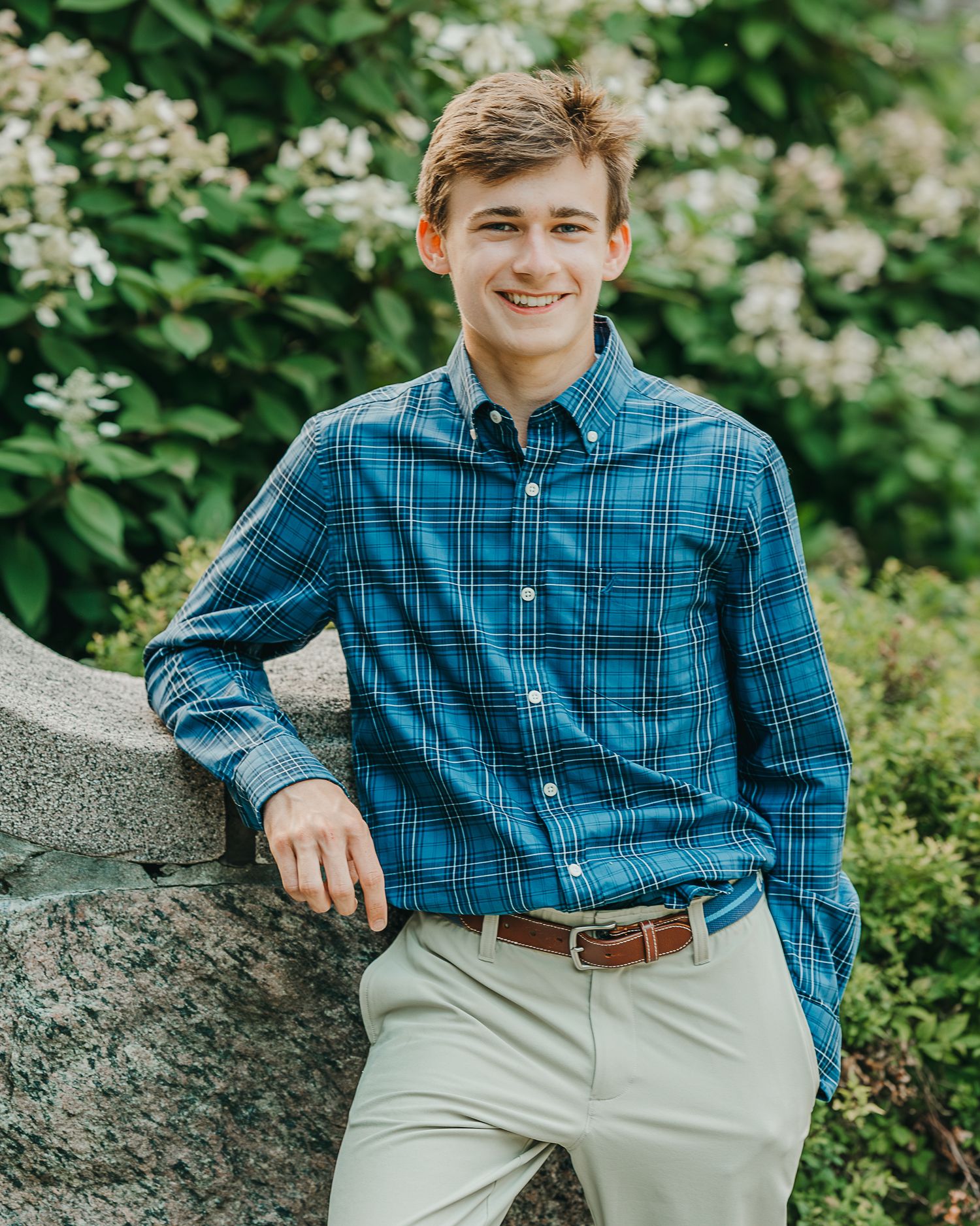 Young man in blue plaid shirt and khaki pants smiling outdoors near stone wall with green foliage background.