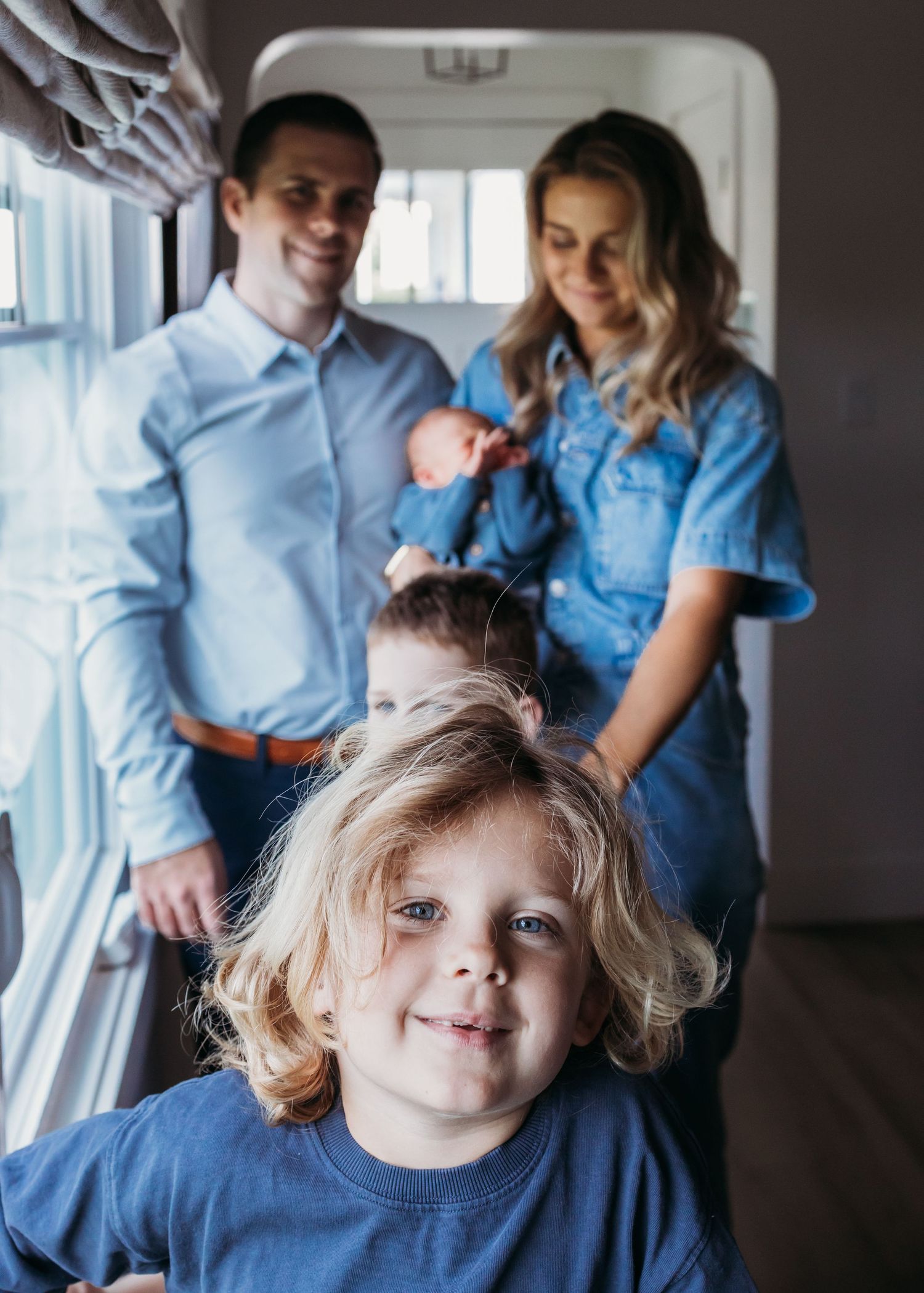 A young family poses indoors, parents holding a newborn while two blond children smile in matching blue outfits.