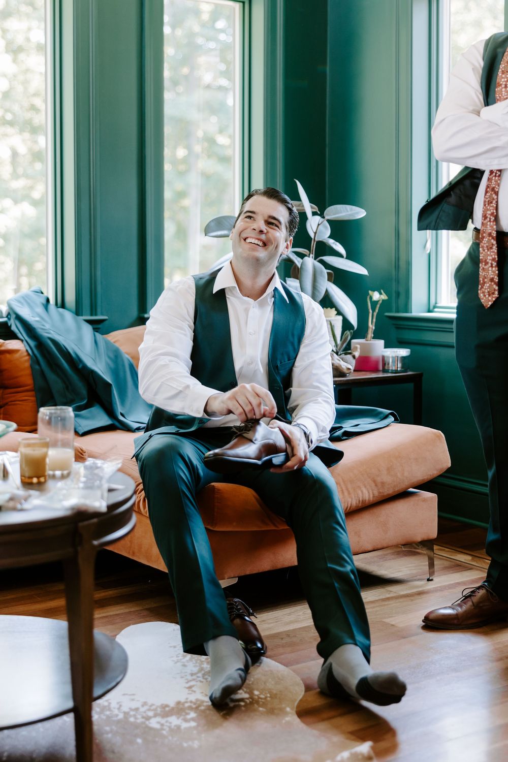 Smiling groom in dark teal suit sits on orange velvet sofa during wedding day preparations indoors.
