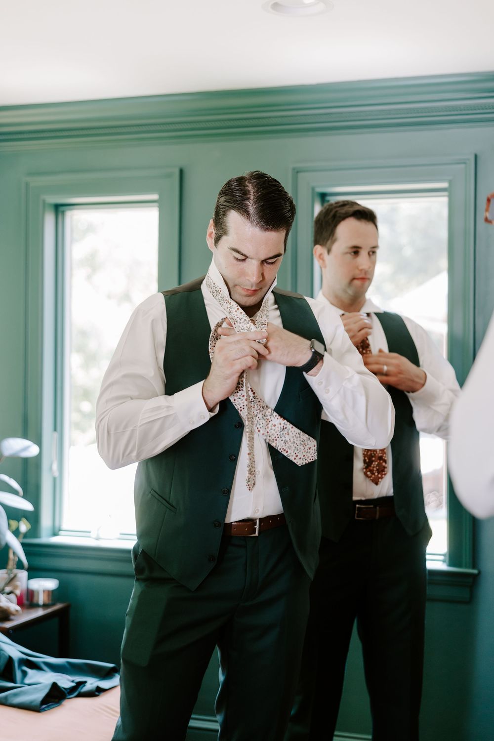 Groom adjusting floral tie in teal room while groomsman ties his own matching necktie nearby.