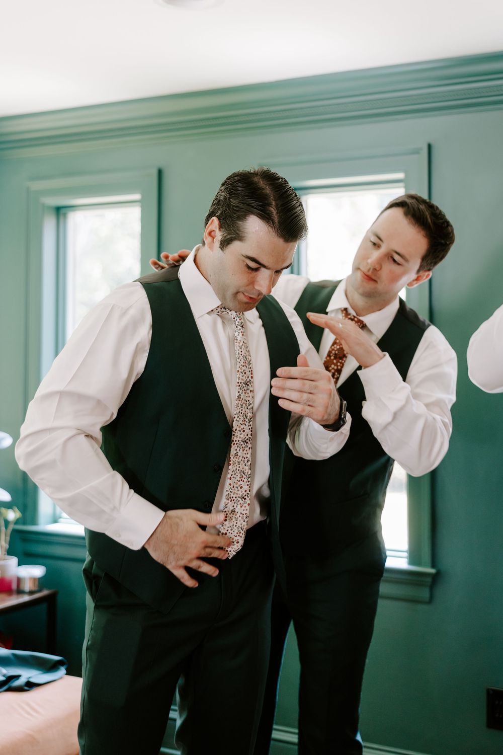 Two groomsmen in dark suits helping each other with floral ties during wedding day preparations.