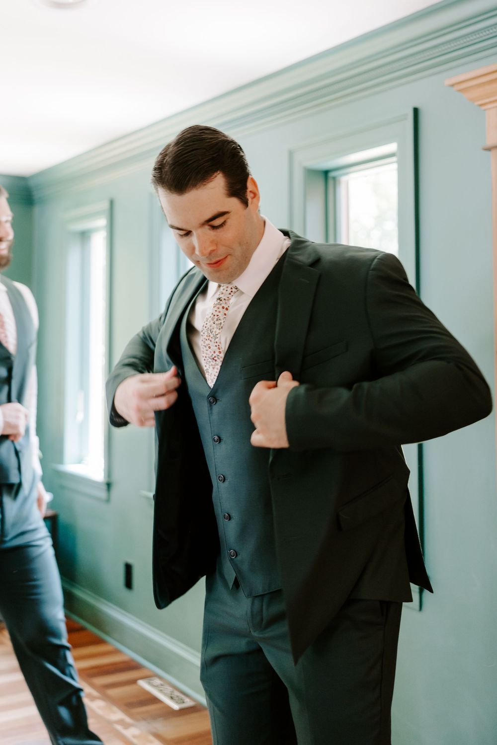 Groom buttoning dark green three-piece suit jacket during wedding day getting ready preparations indoors.