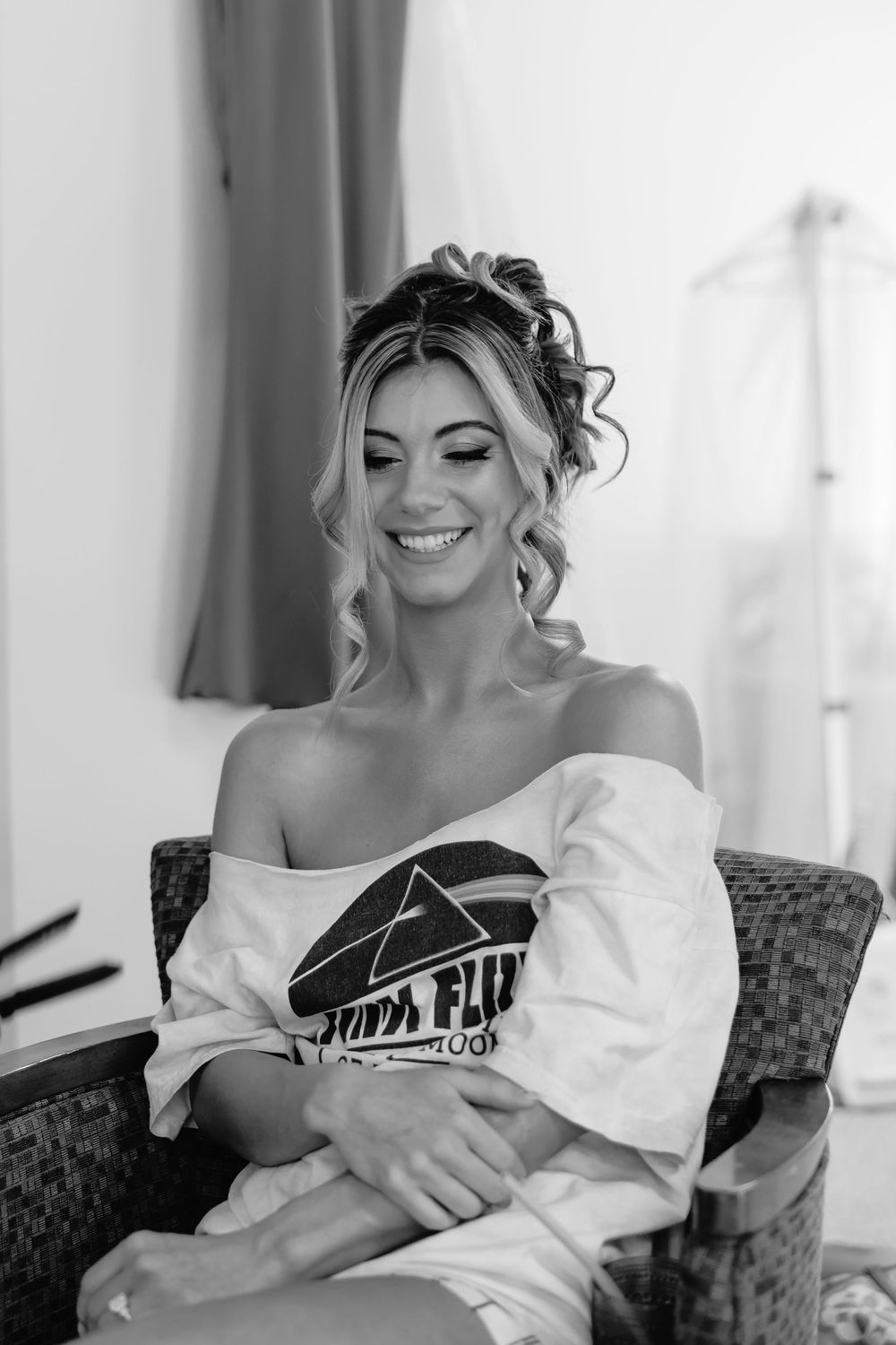 Black and white portrait of radiant bride smiling in off-shoulder shirt during wedding preparations.