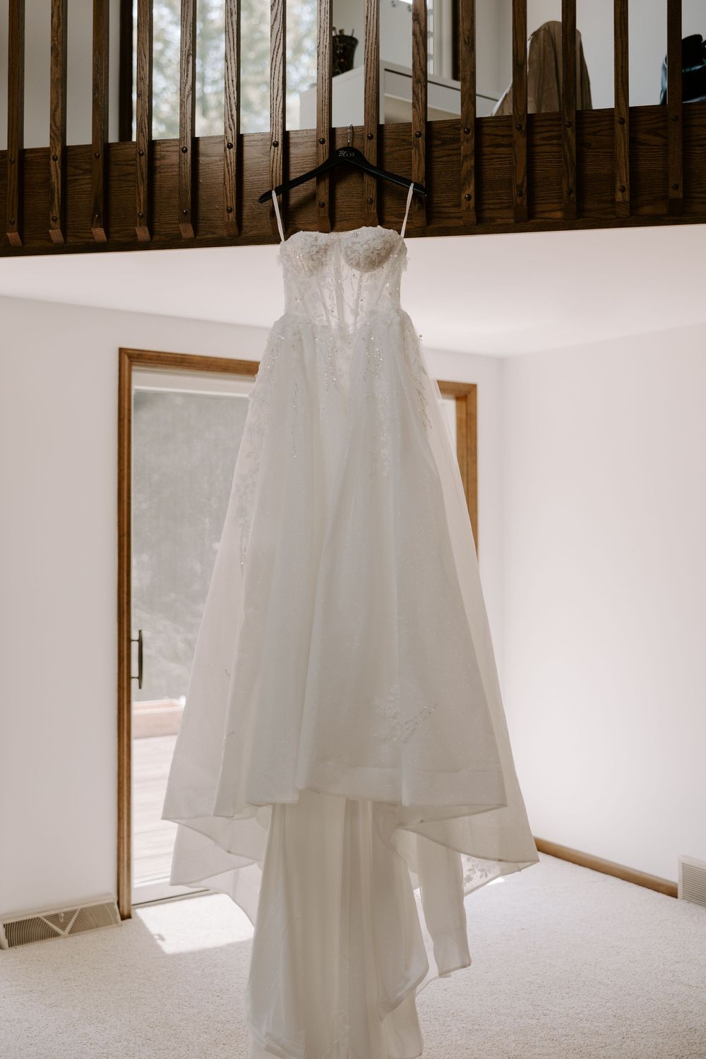 Elegant strapless lace A-line wedding dress hanging from wooden balcony railing in bright room.