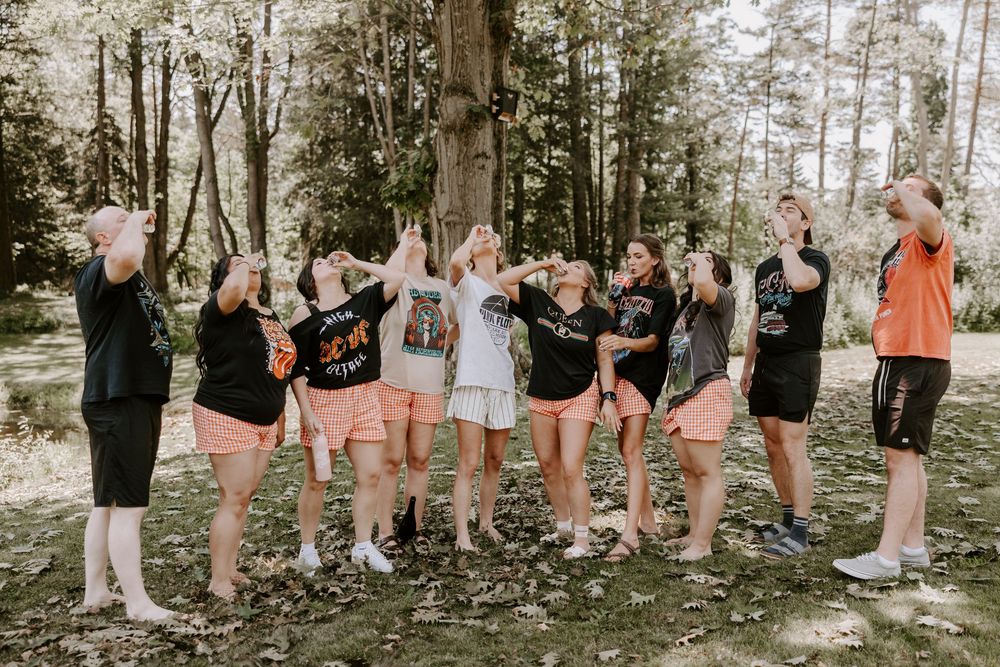 Bridal party in matching black shirts and peach shorts celebrating outdoors among tall trees.