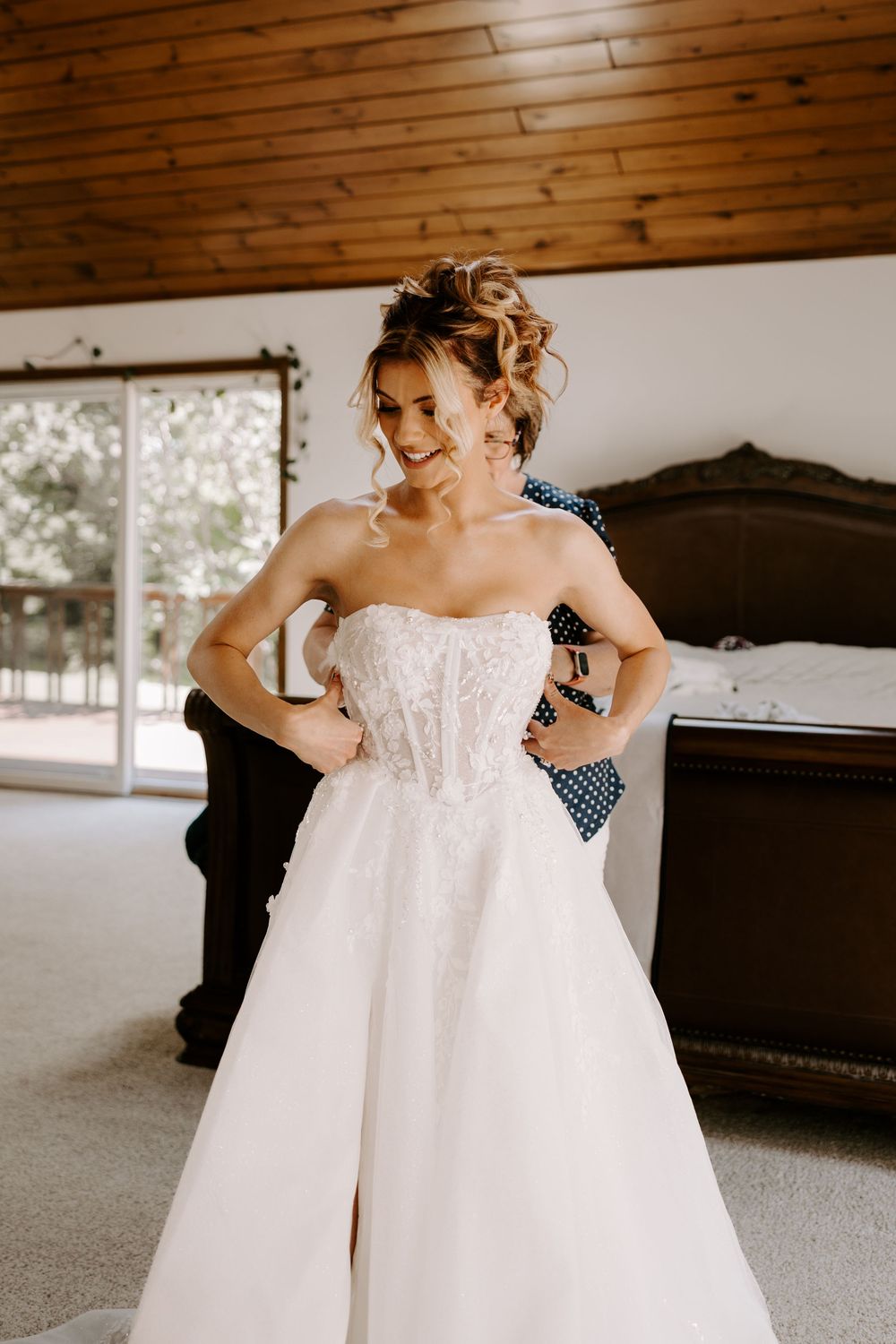 Beautiful bride in strapless lace wedding gown poses hands on hips while being buttoned up.