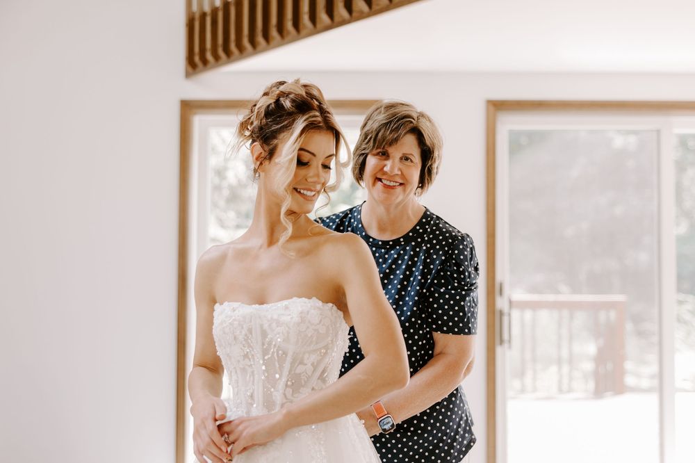 Mother helping bride zip up her strapless lace wedding gown while getting ready indoors.