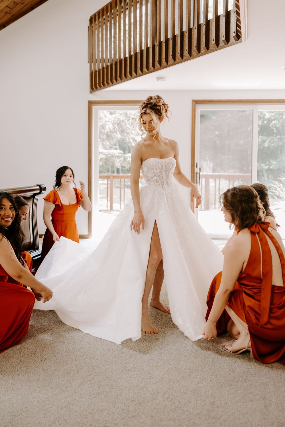 Bridesmaids in rust orange dresses helping bride with her white wedding gown train indoors.