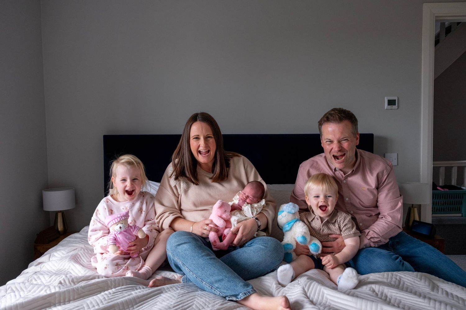 Happy family of five sitting on a bed, parents smiling with three young children including a newborn baby.
