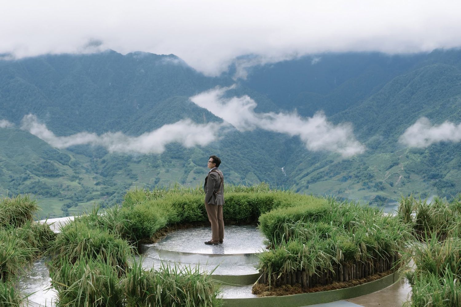 Person standing on terraced rice fields with misty mountain valleys and low clouds in the background.