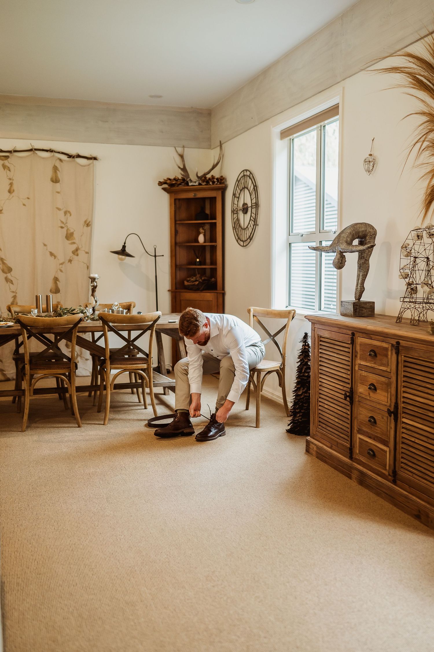 A man in white dress shirt bends down to tie his black shoes in a warmly decorated rustic dining room.