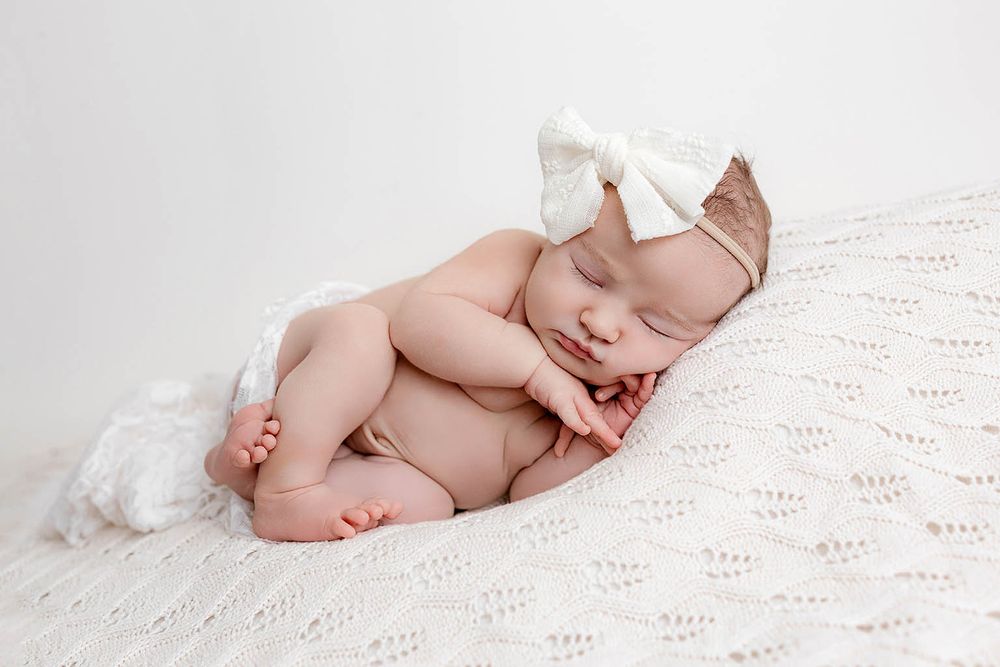Curled sleeping newborn baby with white bow headband resting on soft knit white blanket.