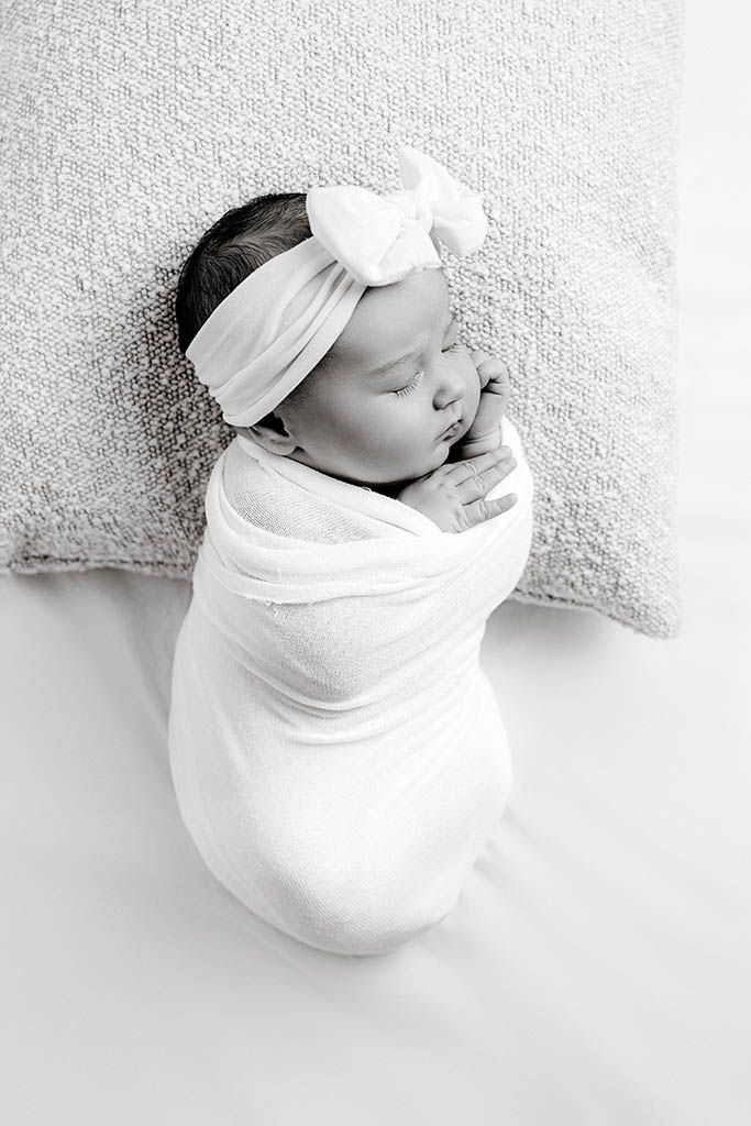 Black and white photo of a sleeping newborn baby swaddled in white wrap with a bow headband on a pillow.