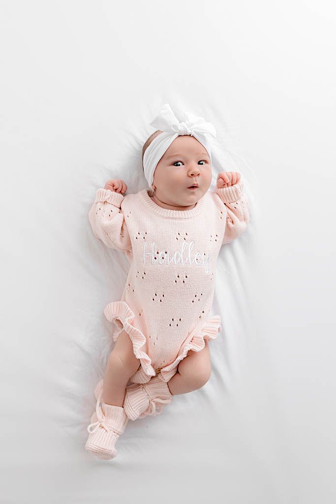 Newborn baby girl in pink knit romper and white bow headband lying on white blanket.