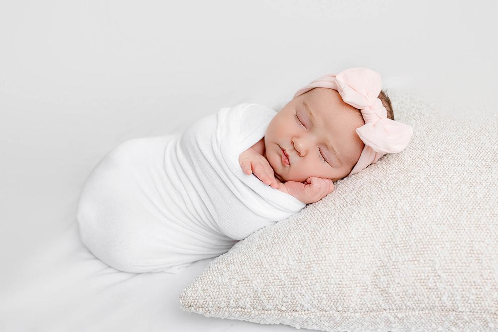 Newborn baby girl swaddled in white wrap with pink bow headband sleeping peacefully on a cream pillow.