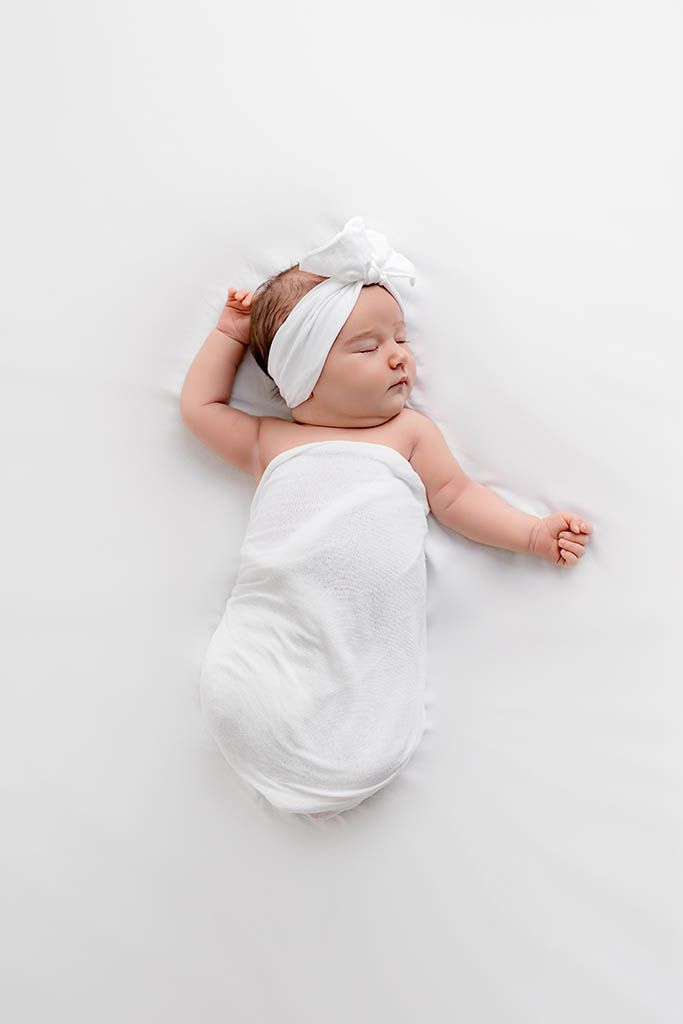Sleeping newborn baby wrapped in white swaddle with bow headband lying on white backdrop.
